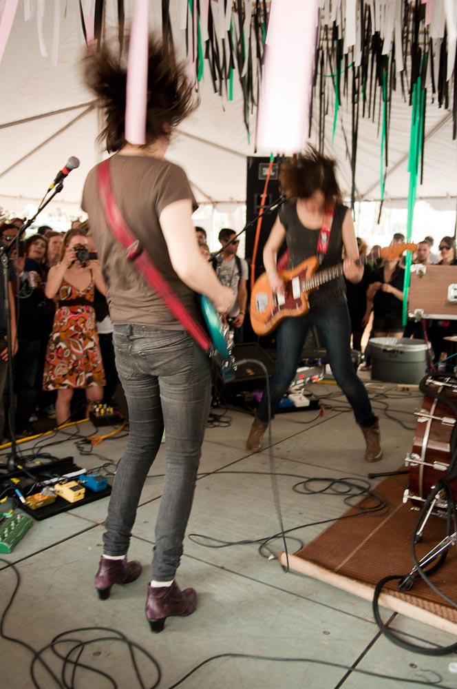 Young fans watch a band of two women play guitar on a stage at a small venue under a large white tent during the day. The image is blurred with the frenetic motion of the bandmates.