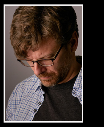 Headshot of Mike Bicknell looking down. He has glasses, a beard, wavy hair, and is wearing a t-shirt and open button down shirt.