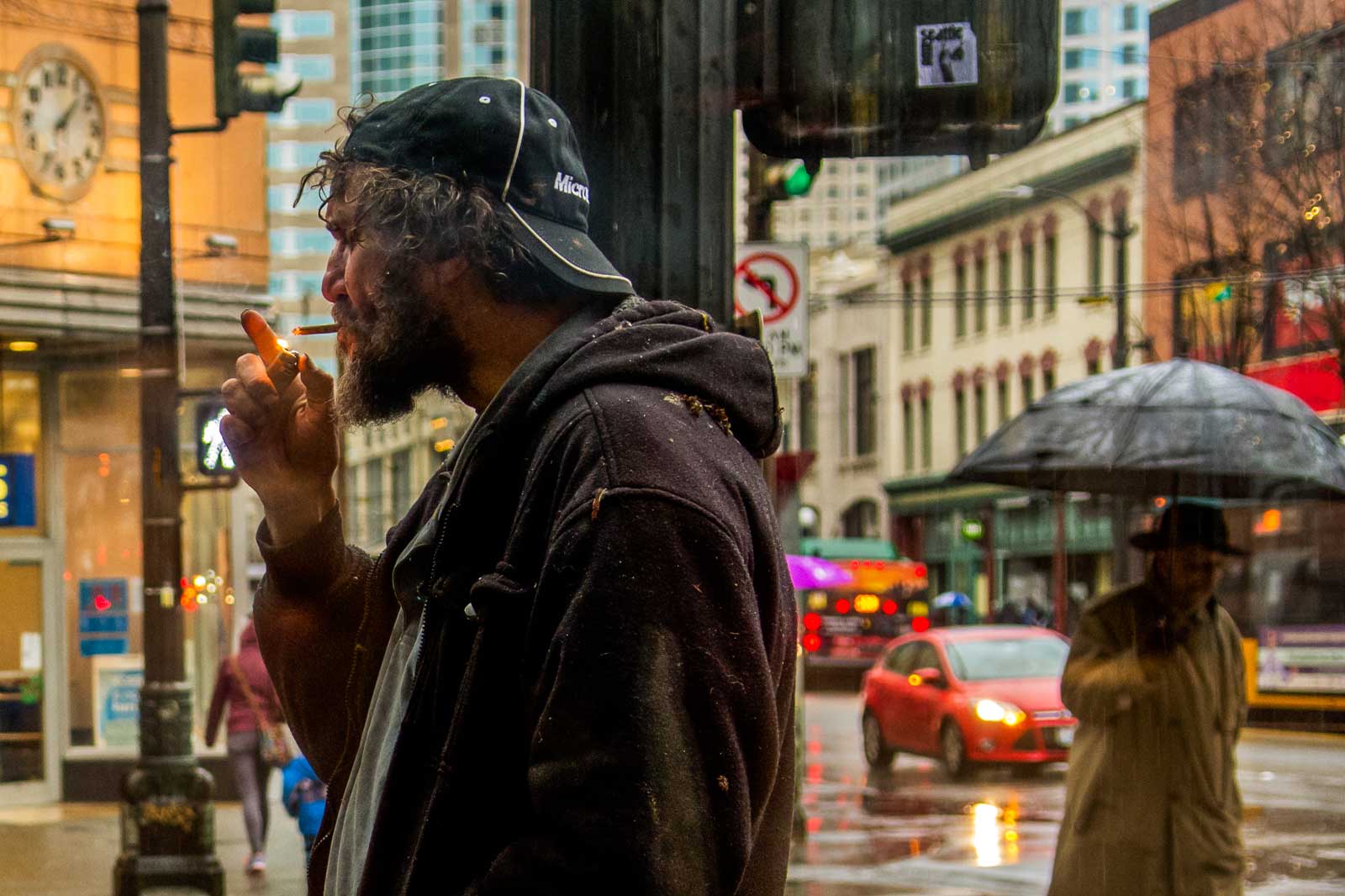 scruffy man lights a cigarette on the sidewalk as people walk and drive by on rainy streets